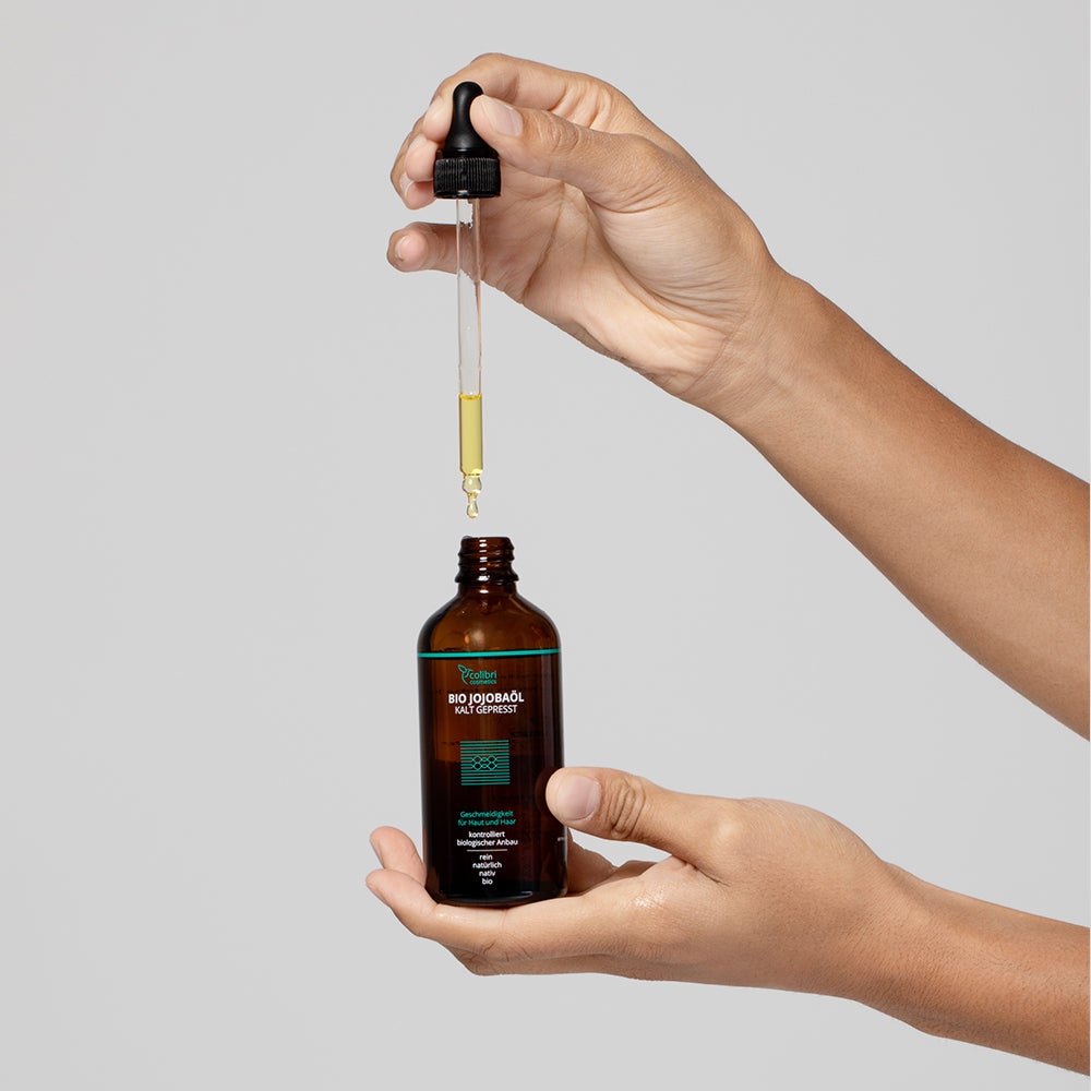 Eine Hand hält eine geöffnete Flasche Jojobaöl mit einer Pipette, aus der Öl tropft.