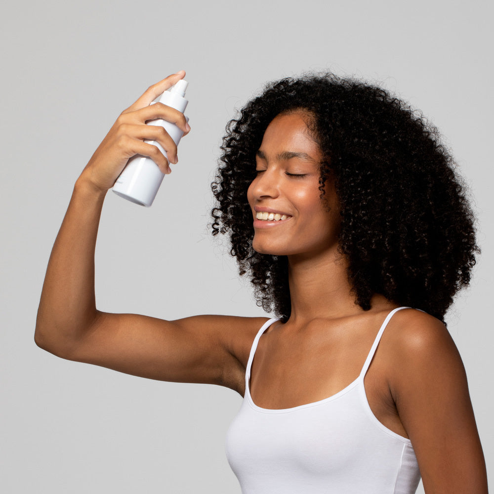Frau mit lockigem Haar sprüht sich lächelnd Wasser oder ein Spray aus einer weißen Flasche ins Gesicht.