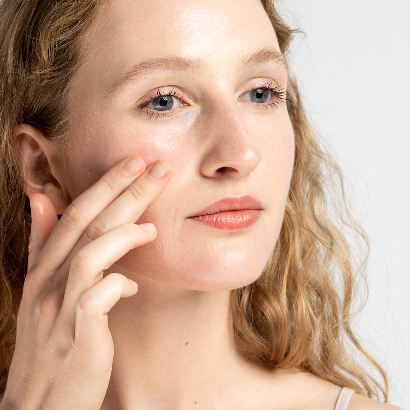 Frau mit lockigem Haar trägt Hautpflegecreme auf ihre Wangen auf und blickt leicht nach oben.