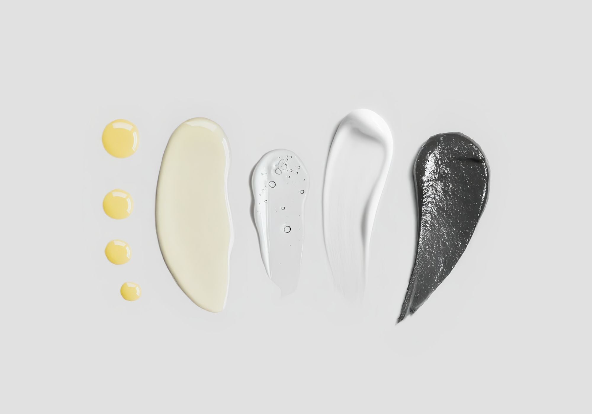 Verschiedene Texturen von Kosmetikprodukten in Gelb, Beige, Transparent, Weiß und Grau auf grauem Hintergrund.