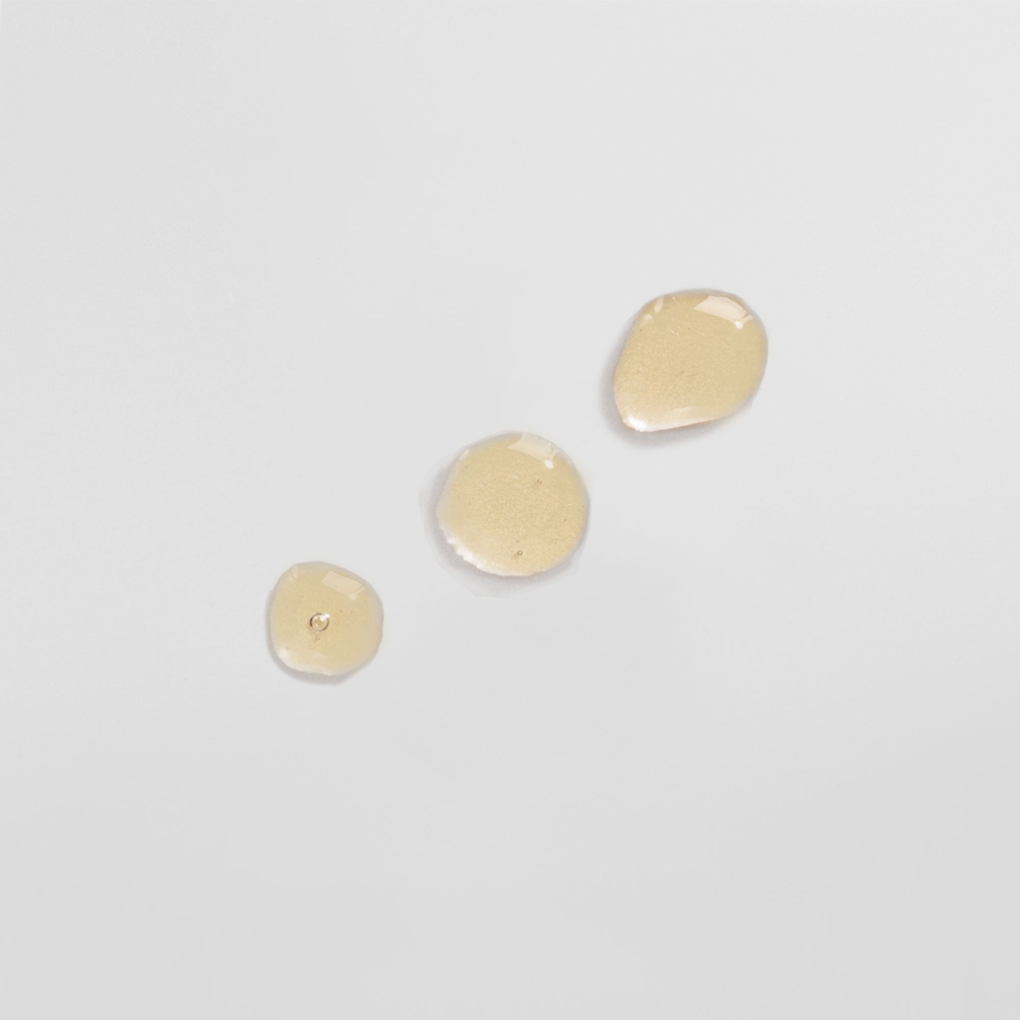 Drei beige Flüssigkeitstropfen auf einer hellen, glatten Oberfläche.