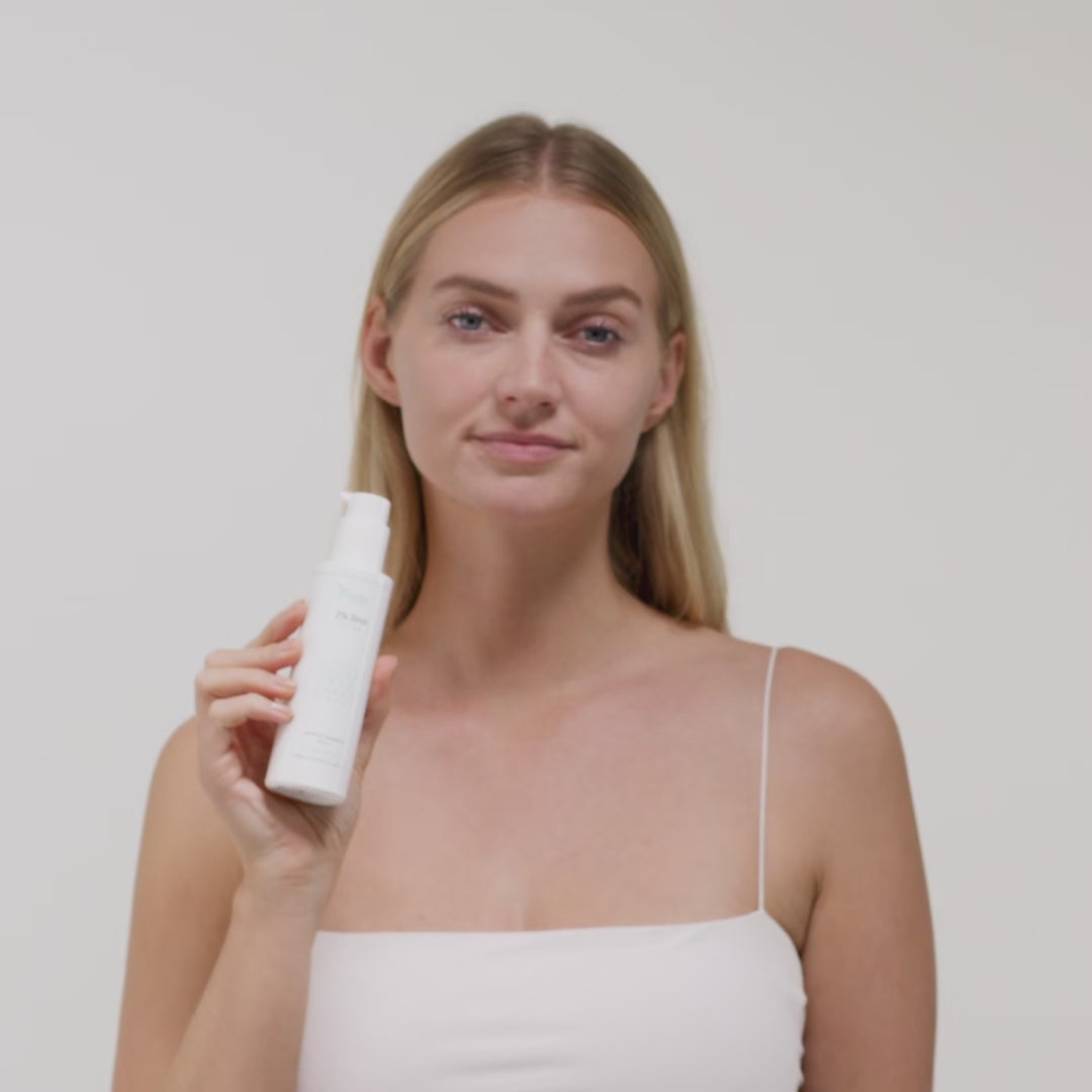 Frau hält eine weiße Flasche mit kosmetischem Produkt vor einem neutralen Hintergrund.