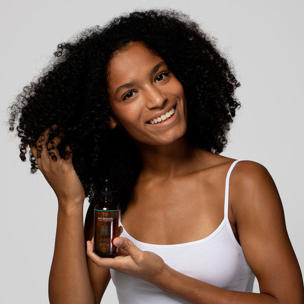 Frau mit lockigem Haar hält eine Flasche Haaröl in der Hand und lächelt in die Kamera.