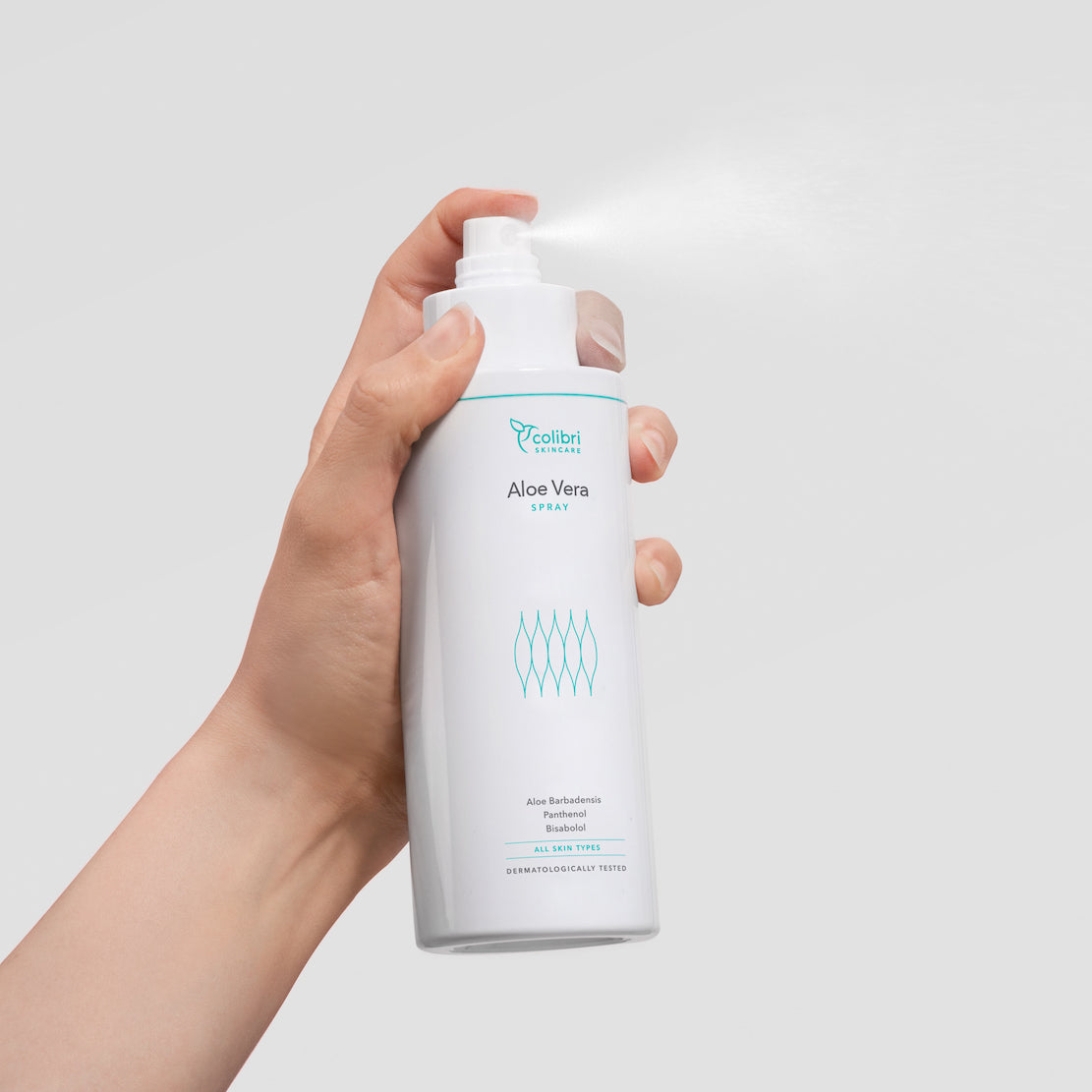 Eine Hand hält eine Flasche Aloe Vera Spray, aus der ein feiner Nebel sprüht, vor einem grauen Hintergrund.