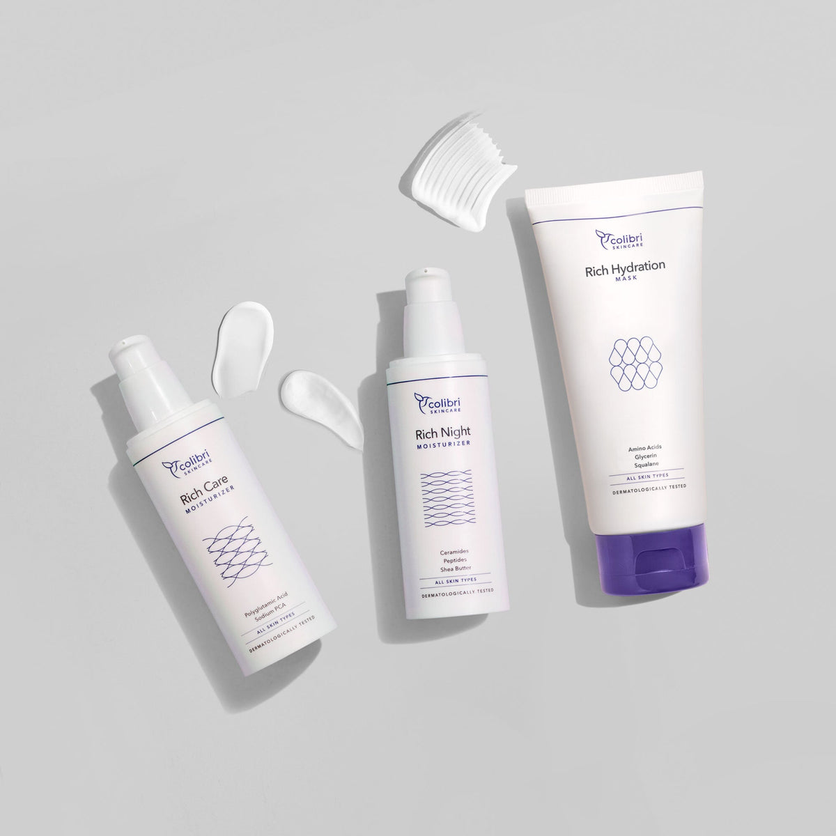 Drei Hautpflegeprodukte auf grauem Hintergrund mit weißen Creme-Tupfern und einer Applikatorbürste.