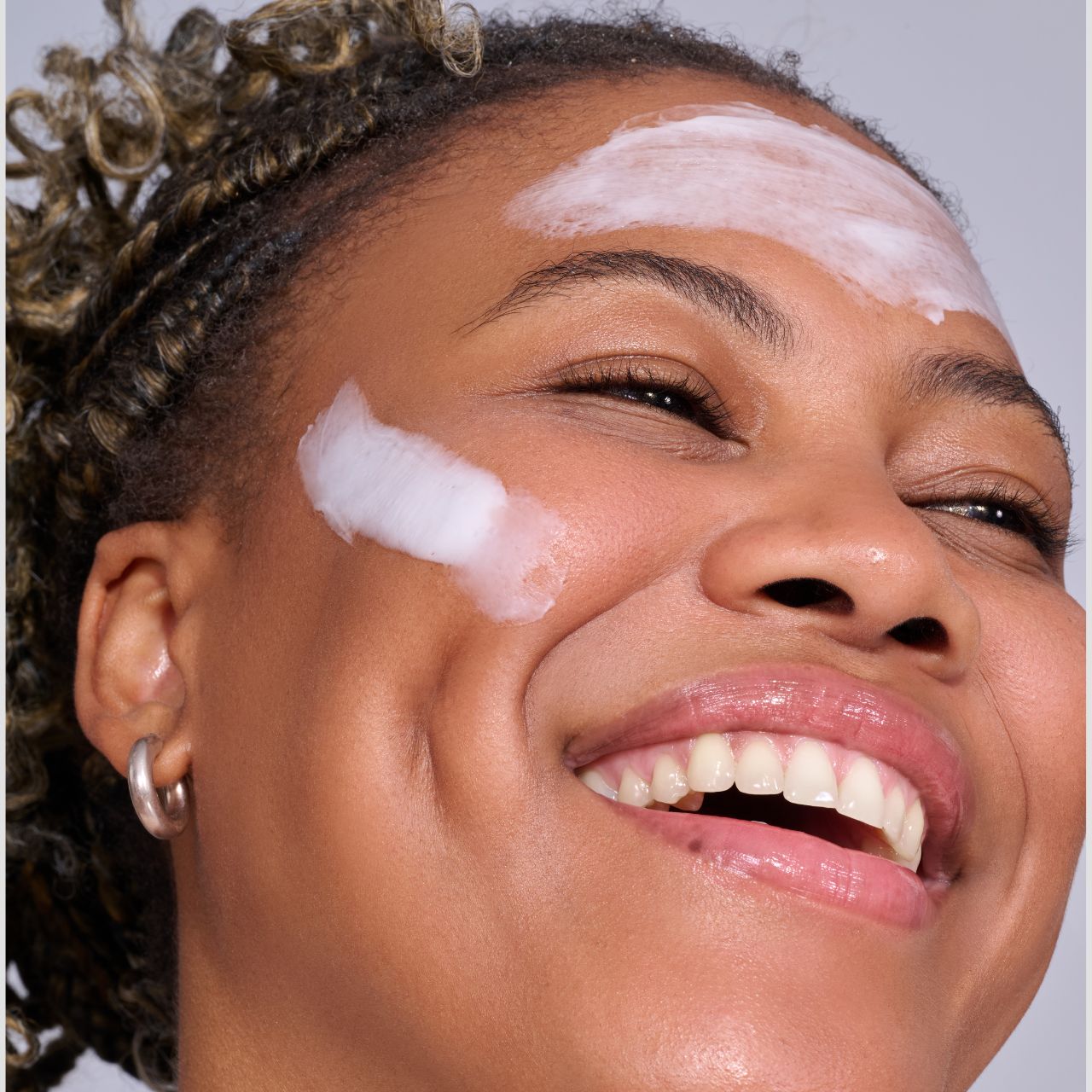 Frau mit strahlendem Lächeln trägt weiße Creme auf Stirn und Wangen auf, Nahaufnahme des Gesichts.