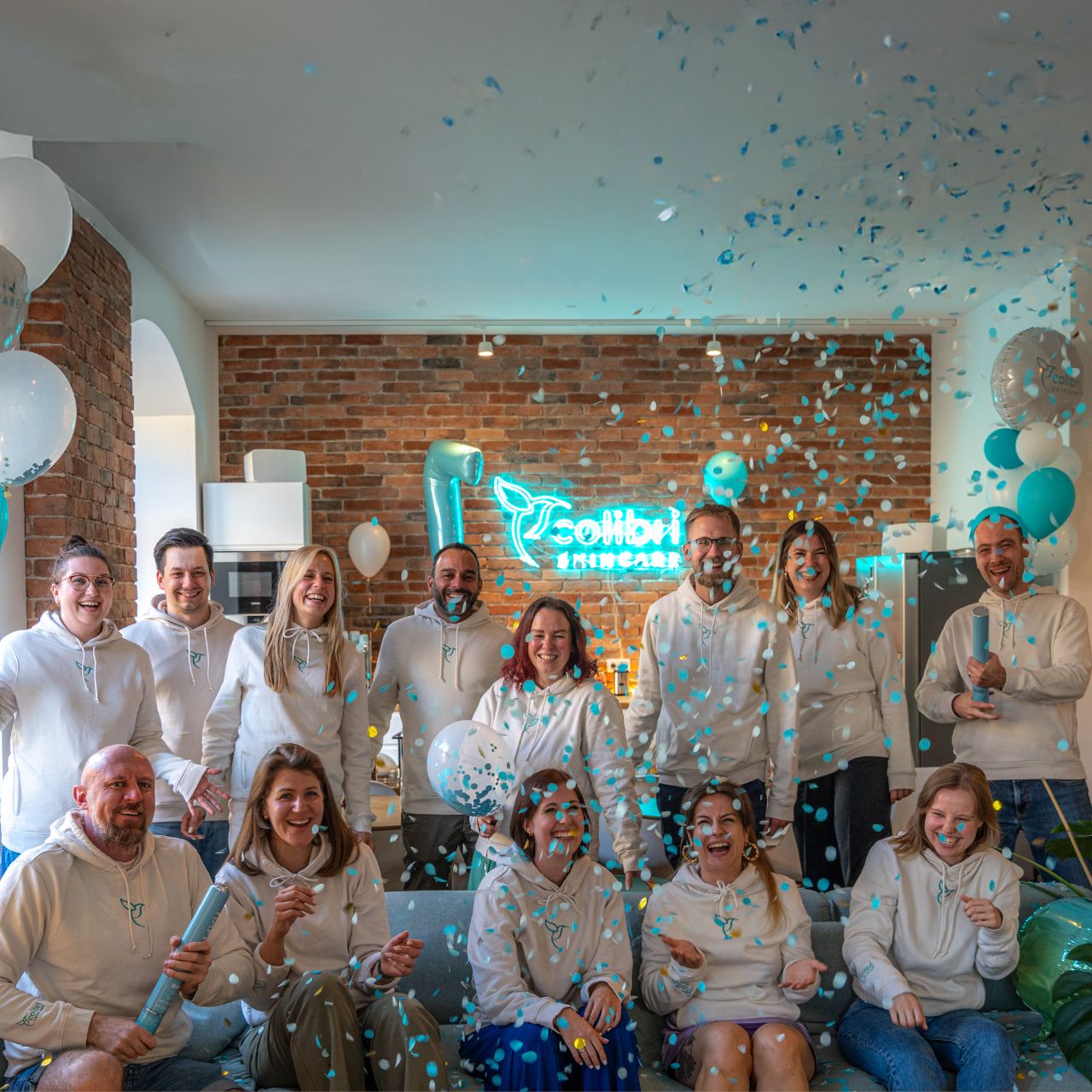 Gruppe Menschen in weißen Hoodies feiert mit Konfetti und Luftballons in einem Raum mit Ziegelwand und Neonlicht.