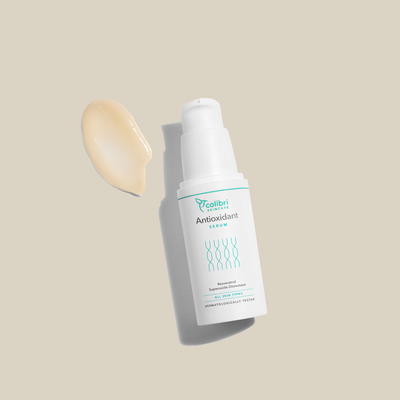 Weißes Serumfläschchen mit "Antioxidant Serum"-Aufschrift, daneben eine beige Cremeprobe, vor beigem Hintergrund.