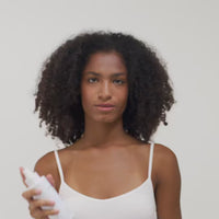 Eine Person mit lockigem Haar hält eine Sprühflasche in der Hand und trägt ein weißes Top vor einem neutralen Hintergrund.